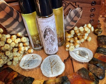May include: Three amber glass bottles with black roll-on tops and labels featuring a woman in a dress and the text "Goddess Magic", "Freyja", and a rune symbol. The bottles are surrounded by amber beads, a feather, and two wooden discs with rune symbols.
