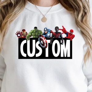 Peut inclure: Sweat-shirt blanc avec un graphique de différents super-héros, dont Iron Man, Hulk, Captain America, Black Panther et Spiderman, avec le mot "CUSTOM" imprimé en dessous.