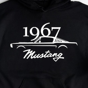Può includere: Felpa con cappuccio nera con grafica bianca di una Mustang del 1967. Il testo "1967" è sopra l'auto, e "Mustang" sotto. La felpa ha una tasca frontale e un cappuccio.