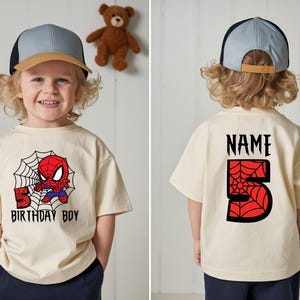 Może przedstawiać: Beżowa koszulka z grafiką Spider-Mana i napisem "BIRTHDAY BOY". Tył koszulki ma napis "NAME" nad czerwoną cyfrą 5 z pajęczyną. Dziecko ma na sobie niebiesko-beżową czapkę z daszkiem.