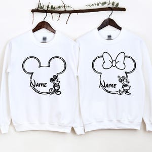 May include: Two white sweatshirts with black outlines of Mickey Mouse and Minnie Mouse heads. The sweatshirts have the word "Name" written inside the outlines.