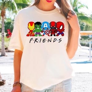 K&ouml;nnte beinhalten: Wei&szlig;es T-Shirt mit einer Grafik von f&uuml;nf Comic-Superhelden, darunter Iron Man, Hulk, Captain America, Black Panther und Spiderman. Der Text "FRIENDS" ist unter der Grafik gedruckt.