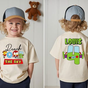 May include: A cream-colored short-sleeve t-shirt featuring a graphic print. The front displays the text Reach Four The Sky with character-themed letters. The back features the name Louis above a large green number four with space-themed details.