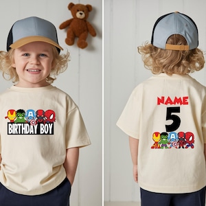 May include: A cream-coloured t-shirt featuring cartoon superhero characters. The front of the shirt says "BIRTHDAY BOY" and the back says "NAME 5". The shirt is worn by a child wearing a blue and tan baseball cap.
