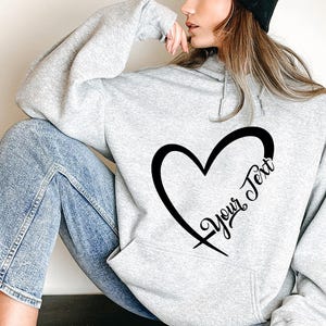 Könnte beinhalten: Ein grauer Hoodie mit einem schwarzen Herzdesign und dem Text "Your Text" im Herzen.