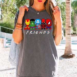 K&ouml;nnte beinhalten: Ein graues T-Shirt mit einem Aufdruck von Comic-Superhelden, darunter Iron Man, Hulk, Captain America, Spiderman und eine schwarze Figur. Der Text "FRIENDS" ist unter dem Aufdruck gedruckt.