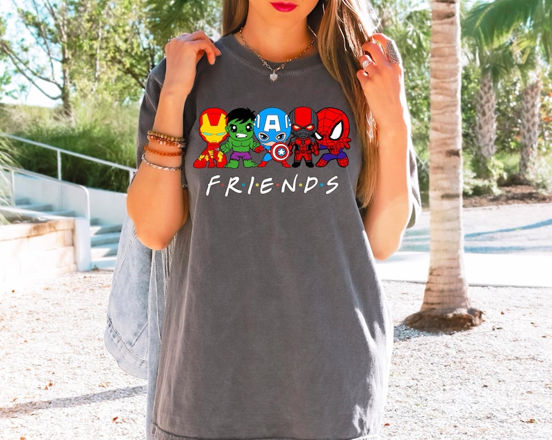 K&ouml;nnte beinhalten: Ein graues T-Shirt mit einem Aufdruck von Comic-Superhelden, darunter Iron Man, Hulk, Captain America, Spiderman und eine schwarze Figur. Der Text "FRIENDS" ist unter dem Aufdruck gedruckt.
