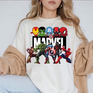 May include: White sweatshirt with a graphic print of various Marvel superheroes, including Iron Man, Hulk, Captain America, Spider-Man, and Thor. The text "MARVEL" is printed in black below the superheroes.