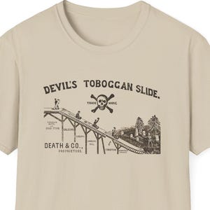 Puede incluir: Una camiseta color beige con un gráfico vintage de una pista de trineo. El gráfico incluye el texto "DEVIL'S TOBOGGAN SLIDE" y una calavera con huesos cruzados. También está presente el texto "DEATH & CO."