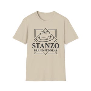 Puede incluir: Camiseta beige con un logotipo en blanco y negro que dice "STANZO BRAND FEDORAS". El logotipo incluye un diseño de sombrero fedora dentro de un marco rectangular. La camiseta tiene mangas cortas y cuello redondo.