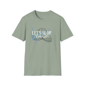 Può includere: T-shirt verde salvia con la scritta "LET'S SLOP 'EM UP!" in bianco, sopra una grafica di roccia con dettagli blu. La maglietta ha maniche corte e girocollo.
