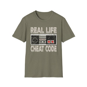 Puede incluir: Camiseta verde oliva con un diseño desgastado de un mando de videojuegos retro. El texto "REAL LIFE CHEAT CODE" está impreso encima y debajo del diseño del mando. La camiseta tiene cuello redondo y mangas cortas.