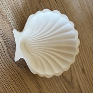 Könnte beinhalten: Eine weiße Keramik-Muschelschüssel. Die Schüssel ist muschelförmig mit einem glatten, abgerundeten Innenraum.