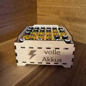 Könnte beinhalten: Eine Holzkiste gefüllt mit AA-Batterien. Die Kiste hat einen Blitz und die Worte "volle Akkus" auf der Seite eingraviert. Die Batterien haben goldene und schwarze Oberteile. Die Kiste steht auf einer Holzoberfläche.