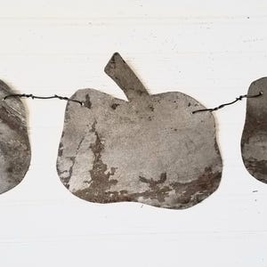 May include: A rustic metal apple garland with three weathered apple shapes strung on a wire. The apples have a distressed, aged appearance with visible rust and imperfections, creating a vintage aesthetic. The garland is suitable for fall or farmhouse decor.