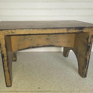 May include: A small, rectangular wooden step stool with a distressed finish. The stool has a dark brown top and a tan base with black accents. The legs are slightly curved, and the overall design is rustic.