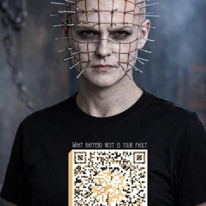 May include: A person with a bald head, adorned with nails and a red grid, wearing a black t-shirt. The shirt features a QR code design with the text "WHAT HAPPENS NEXT IS YOUR FAULT". The background is a dark, smoky gray.