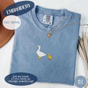 Könnte beinhalten: Ein blaues Jeans-T-Shirt mit einem gestickten Design einer weißen Ente und einem gelben Entlein. Das Shirt hat ein Comfort Colors-Etikett und eine goldene Halskette. Der Text "EMBROIDERY" und "Let me know if you want to change something!" sind sichtbar.