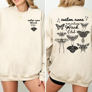 May include: A sand-colored crewneck sweatshirt featuring black ink designs. The front displays the text custom name book club with a moth graphic. The back features the text custom name romantasy book club surrounded by various moth illustrations.
