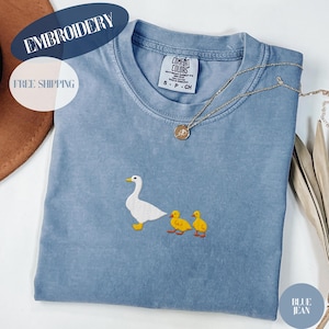 May include: A blue jean colored t-shirt with an embroidered design of a white duck and two yellow ducklings. The shirt has a crew neck and short sleeves. A gold necklace with a pendant is draped on the shirt.