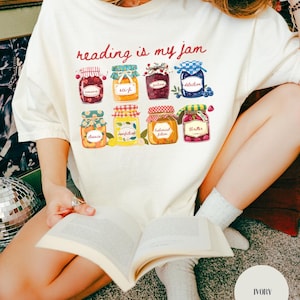 Può includere: T-shirt color crema con la scritta "reading is my jam" e illustrazioni di vasetti di marmellata. I vasetti sono etichettati con generi di libri come romance, fantascienza e fantasy. La persona tiene in mano un libro.