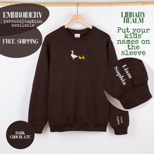 Könnte beinhalten: Dunkelbraunes Sweatshirt mit gesticktem Design einer weißen Gans und zwei gelben Entenküken. Am Ärmelbündchen stehen die Namen "Liam" und "Sophia" in Weiß. Der Text auf dem Bild lautet "STICKEREI Personalisierung verfügbar" und "Namen Ihrer Kinder auf den Ärmel schreiben."