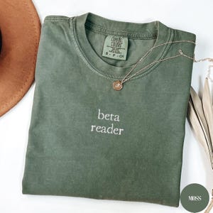May include: An olive green t-shirt with the embroidered text "beta reader". The shirt has a crew neck and short sleeves. A gold necklace with a pendant is draped across the shirt. The shirt is labelled "Comfort Colors".