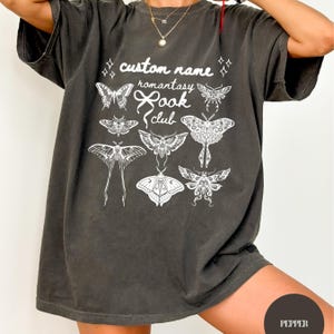 May include: A charcoal gray short-sleeve t-shirt featuring a white graphic print of various moth and butterfly illustrations. The text reads custom name romantasy book club, accompanied by a bow icon and small star accents on the front of the shirt.