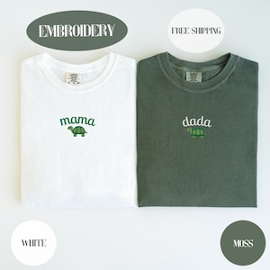 May include: Two crewneck t-shirts, one white and one moss green. The white shirt features green embroidered text reading mama above a small turtle graphic. The moss green shirt features white embroidered text reading dada above a small turtle graphic.