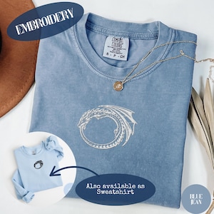 以下が含まれることがあります： 青いジーンズ色のTシャツで、白い刺繍のドラゴンが円形にデザインされています。首元にはComfort Colorsのラベルが付いています。小さな円形のペンダントネックレスが生地の上に置かれています。円形のインセットにはスウェットシャツが表示されています。
