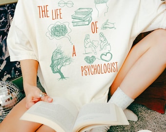 May include: Ivory t-shirt with the text "THE LIFE OF A PSYCHOLOGIST" in red and green. The shirt features illustrations of books, a brain, and a tree. The person is holding an open book.
