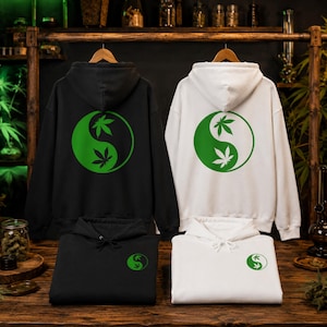 Könnte beinhalten: Zwei Kapuzenpullover, einer in Schwarz und einer in Weiß, jeweils mit einem großen grünen Yin-Yang-Symbol auf der Rückseite, das Cannabisblatt-Silhouetten enthält. Kleinere Versionen desselben grünen Yin-Yang-Blatt-Motivs sind vorne aufgedruckt.