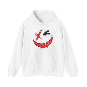 Puede incluir: Una sudadera con capucha blanca que presenta un estampado gr&aacute;fico en el pecho. El dise&ntilde;o muestra un rostro estilizado con un ojo rojo tachado, un ojo negro s&oacute;lido y una boca roja ancha y dentada con dientes afilados y efectos de pintura que gotea.