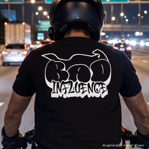 May include: A black short-sleeved t-shirt featuring a large white graphic on the back. The design includes a stylized motorcycle silhouette above the bold, bubble-lettered text "BAD" and the word "INFLUENCE" written in a graffiti-style font.