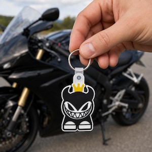 May include: A black and white rubber keychain featuring a stylized character with horns and a wide, toothy grin. The character wears a small yellow crown on its head. The keychain is attached to a silver metal ring and a translucent plastic loop.