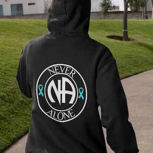 Pode incluir: Moletom preto com capuz com um design circular branco nas costas. O design inclui o texto "NEVER ALONE" e as letras "NA" no centro, com fitas turquesas em ambos os lados.