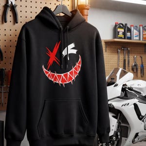 Puede incluir: Una sudadera negra con capucha y un estampado gr&aacute;fico en el pecho. El dise&ntilde;o muestra un rostro estilizado con una X roja sobre un ojo, un parche blanco sobre el otro y una amplia sonrisa dentada de color rojo y blanco con dientes afilados.