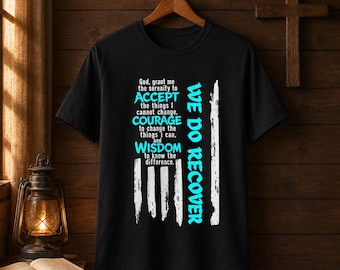 Pode incluir: Uma t-shirt preta de manga curta com um design gráfico de uma bandeira americana branca desgastada. O texto diz: God, grant me the serenity to ACCEPT the things I cannot change, COURAGE to change the things I can, and WISDOM to know the difference. WE DO RECOVER em azul brilhante.
