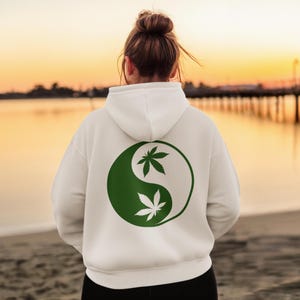 Könnte beinhalten: Weißer Kapuzenpullover mit einem grün-weißen Yin-Yang-Design mit Cannabisblättern. Der Hoodie hat eine Kapuze mit Kordelzug und eine Kängurutasche. Das Design ist auf der Rückseite zentriert. Der Hintergrund zeigt einen Strand und einen Pier bei Sonnenuntergang.