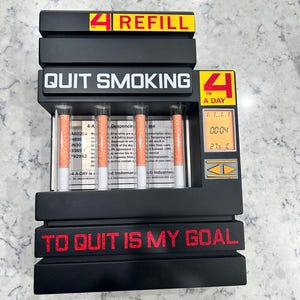 Może przedstawiać: Czarny, prostokątny dozownik do papierosów z napisami 4 REFILL, QUIT SMOKING oraz 4 A DAY. Posiada cztery pionowe szklane rurki z papierosami, cyfrowy wyświetlacz pokazujący 00:04 i 27,5 C oraz czerwony napis TO QUIT IS MY GOAL.