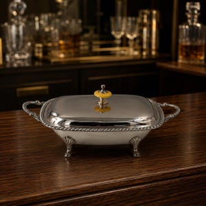 May include: A silver-toned rectangular serving dish with a matching lid, featuring ornate handles, a decorative rope-style border, and four shell-shaped feet. A small, circular yellow knob sits atop the center of the lid for easy lifting.