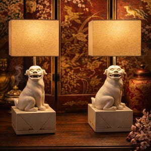 May include: Two white ceramic dog-shaped table lamps with rectangular beige shades. The lamps sit on square bases and are placed on a dark wooden surface. The background features ornate red and gold Asian-inspired decor.