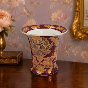 May include: A decorative vase with a wide, fluted rim and a bulbous base. The vase is a deep burgundy color with gold floral designs. The interior is white. The vase is likely made of ceramic or porcelain.