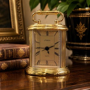May include: A gold-toned rectangular carriage clock with a polished metal finish, featuring a handle on top, a gold face with black Roman numerals, and the text LINDEN and QUARTZ printed on the dial. The clock has three black hands.