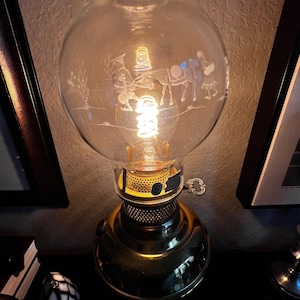 May include: Vintage brass oil lamp with a clear glass globe. The globe features an etched scene of figures with a donkey. The lamp has a round base and a key for adjusting the flame.