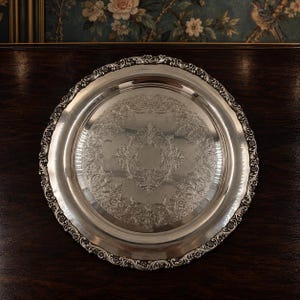 May include: A round silver-toned serving tray featuring an intricate embossed floral and scrollwork pattern in the center. The rim of the tray is decorated with a detailed scalloped edge consisting of repeating floral motifs and curved lines.