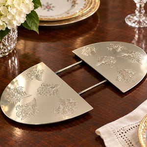 May include: A silver-toned metal trivet featuring two semi-circular plates connected by two parallel rods. Each semi-circular section is decorated with an embossed floral pattern. The metallic surface is reflective and smooth.