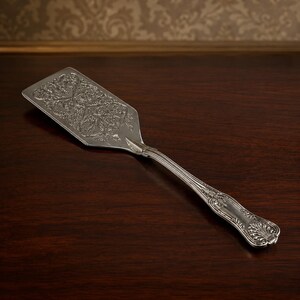 May include: A silver-toned metal cake server featuring an elongated, flat, rectangular blade with intricate, embossed floral and scrollwork patterns. The handle is slender and curved, decorated with detailed engravings and a shell-like motif at the end.