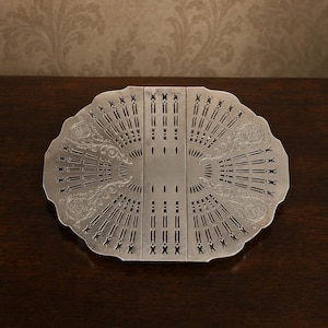 May include: A silver-toned metal trivet featuring a scalloped edge and an intricate, symmetrical cut-out pattern. The design includes two mirrored sections with delicate engraved floral scrollwork flanking a central vertical panel with slots.
