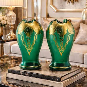 May include: Two green glass vases with gold floral designs. The vases have a unique shape with a ruffled top and a rounded base. The gold detailing includes leaf and vine patterns. The vases are displayed on a stack of books.